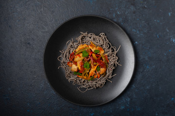 Wheat noodles with black sesame, fried in a wok with chicken and vegetables. Top view. Served on a black plate.