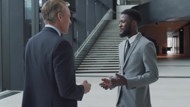 Thigh-up arc shot of two cheerful Caucasian and Afro-American businessmen in suits and ties standing in office hallway near entrance, having friendly conversation, smiling and gesticulating