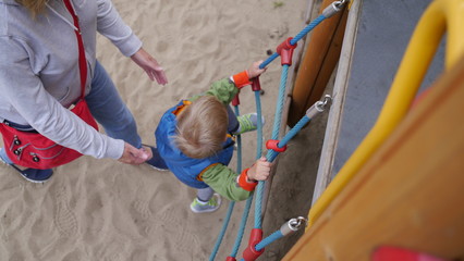 Dziecko wspina się po linach na placu zabaw, mama pomaga mu i podtrzymuje, żeby nie spadł. - obrazy, fototapety, plakaty