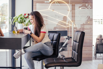 Positive cheerful young girl student communicates in social networks using a smartphone and wireless internet and drinks hot cocoa sit at a table in a cafe. Cozy cafeteria, people lifestyle