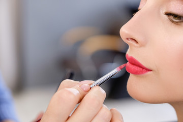 Hand of a professional make-up artist's master dyes the red lips of a young blonde client girl while creating makeup for a photo shoot in beauty room or luxury spa salon. Fashion shooting concept