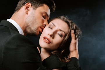 handsome man kissing young woman with closed eyes on black with smoke