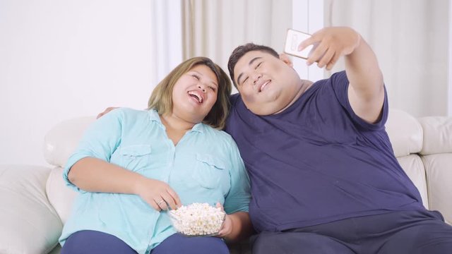 Happy overweight couple using a mobile phone to take selfie photo together while sitting on the sofa in the living room at home. Shot in 4k resolution