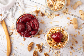 Jar of homemade granola with yogurt and canned strawberry on white wooden background top view