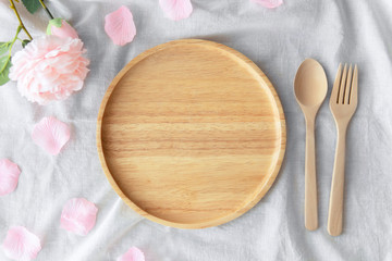 Top view above empty bamboo light brown wooden round plate next to spoon and fork decoration with artificial soft light pink rose flower surrounded by rose petals on crumpled white color fabric cloth.
