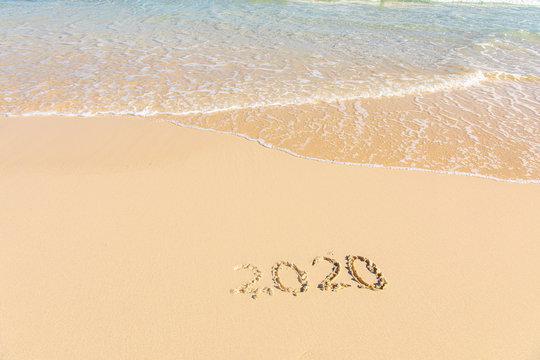 Summer beach holiday 2020 season golden sand - year - decade - new season lettering on the beach with wave and clear blue sea. Numbers 2020 year on the sea shore, message handwritten New Years concept