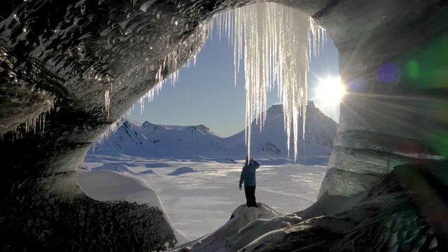 Aerial dolly from inside cave towards snow-clad sunlit mountain range. Explorer views snowy plain of Myrdalsjokull from ice grotto.