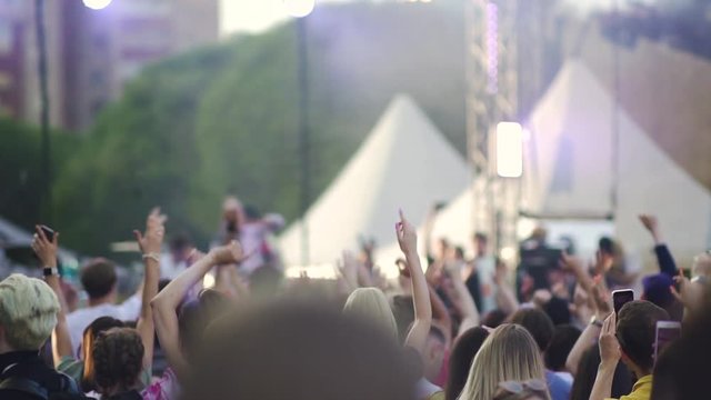 Young people dancing and having fun at music festival on summer evening outdoors. American fans of men and women are enjoying and taking picture holding smartphones in hands in front of stage with