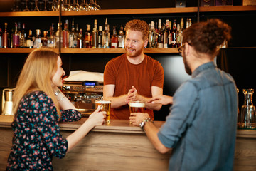 Cheerful friends leaning on bar counter, drinking beer and chatting with bartender. Night out.