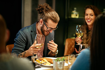 Young handsome caucasian hipster sitting with friends in restaurant, holding glass of wine and laughing.