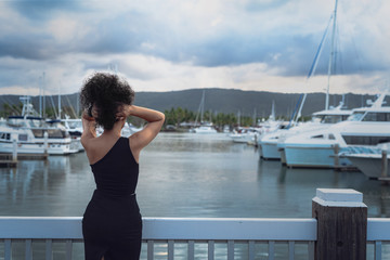 A beautiful young curly-hair woman in a little black dress is posing in the marina. Portrait Photography. Port Douglas, QLD, Australia.