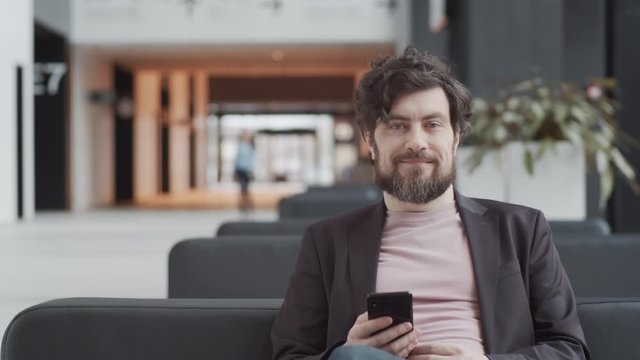 Waist-up portrait shot of handsome middle-aged Caucasian man with full beard, in t-shirt and blazer sitting on couch in departure lounge at airport, holding smartphone, looking at camera and smiling