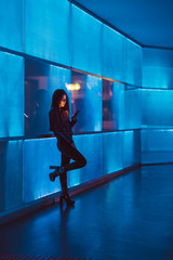 Fototapeta na wymiar Attractive young long-haired girl with glasses looking at her smartphone or mobile telephone leaning next to neon panel with blue light. The color of the lights is blue