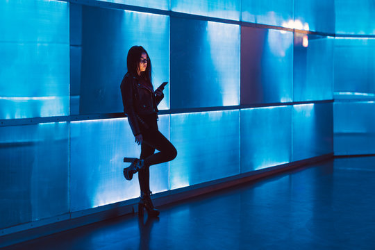 Attractive young long-haired girl with glasses looking at her smartphone or mobile telephone leaning next to neon panel with blue light. The color of the lights is blue