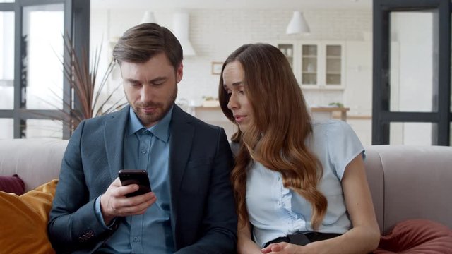 Business couple relaxing on couch together. Annoyed woman looking at man phone