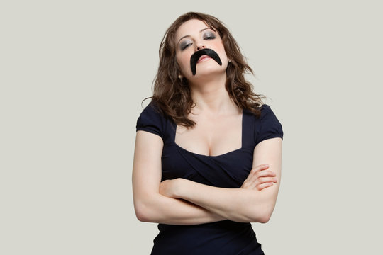 Young woman with arms crossed wearing fake mustache over gray background