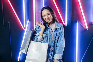 Young girl in blue denim jacket with shopping bags on neon lamps background.