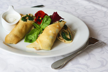 Thin crepes stuffed with spinach and mushrooms. With basil, arugula, violet flowers and sour cream. The dish is served on a table covered with a white tablecloth.