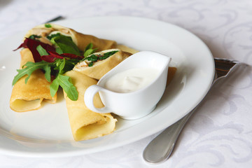 Thin crepes stuffed with spinach and mushrooms. With basil, arugula, violet flowers and sour cream. The dish is served on a table covered with a white tablecloth.