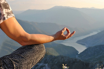 Matured woman in casual clothing practicing yoga outdoor; leg and hand of athlete woman sitting in lotus pose on the top of mountain.