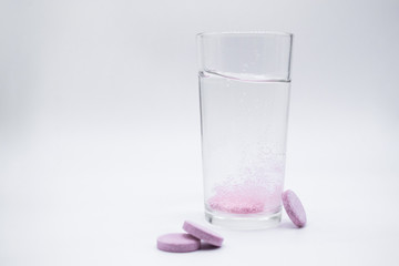 In a clear glass glass with clear water, a soluble,. instant or dissolve pink tablet bubbles. Nearby are similar tablets: vitamin C, dietary supplements, painkillers. White background, copy space