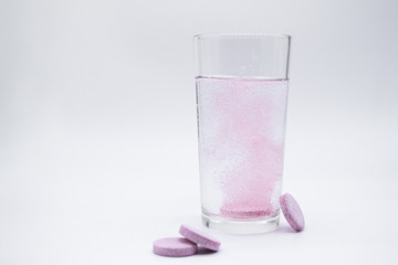 In a clear glass glass with clear water, a soluble,. instant or dissolve pink tablet bubbles. Nearby are similar tablets: vitamin C, dietary supplements, painkillers. White background, copy space