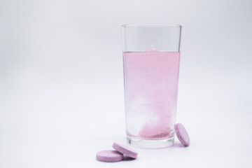 In a glass with water, a soluble, instant or dissolve pink tablet bubbles. Nearby are similar tablets: vitamin C, dietary supplements, painkillers. White background, copy space