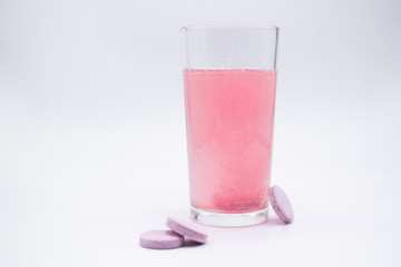 In a glass with pink water, a soluble, instant or dissolve pink tablet bubbles. Nearby are similar tablets: vitamin C, dietary supplements, painkillers. White background, copy space