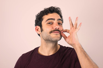 Brown, smiling, handsome man touching his mustache with purple t-shirt. Funny portrait. Pink background.