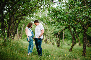 Naklejka na ściany i meble Pregnant girl and her husband are happy to hold hands, stand in the outdoor in the garden background with trees. full length. Looking down at the stomach.