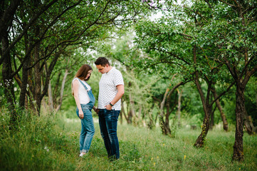 Pregnant girl and her husband are happy to hold hands,  stand in the outdoor in the garden background with trees. full length. Looking down at the stomach.