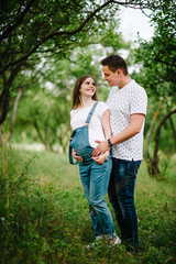 Fototapeta premium Pregnant girl and her husband are happy to hug, hold hands on stomach, stand on grass in the outdoor in the garden background with trees. Close up. full length. Look at each other.