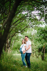 Fototapeta premium Pregnant girl and her husband are happy to hug, hold hands on stomach, stand on grass in the outdoor in the garden background with trees. Close up. full length. Looking at camera