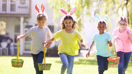 Group Of Children Wearing Bunny Ears Running To Pick Up Chocolate Egg On Easter Egg Hunt In Garden