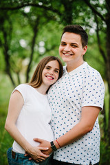 Fototapeta premium Pregnant girl and her husband are happy to hug, hold hands on stomach, stand in the outdoor in the garden background green. Close up. upper half. looking at camera