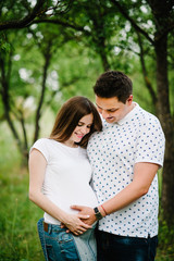 Pregnant girl and her husband are happy to hug, hold hands on stomach,  stand in the outdoor in the garden background green. Close up. upper half. looking  down