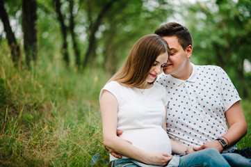 Pregnant girl and her husband are happy to hug, hold hands on stomach, sitting on the grass in the outdoor in the garden background. Close up. upper half. Looking down