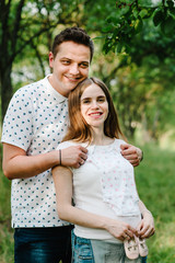 Pregnant girl and hands husband hold hug, to put baby shoes and clothes on stomach, waiting baby. stand in the outdoor in the garden background. Close up. upper half. looking at camera