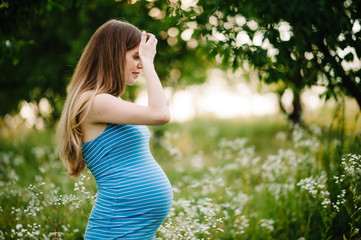 Naklejka na ściany i meble Pregnant serious girl stand and hold hands on head, stand in the outdoor in the garden background with green trees. Close up. upper half. half length. Looking down, sideways.