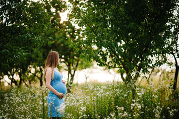 Pregnant happy girl stand and hold hands on stomach,  stand in the outdoor in the garden background with green trees. upper half. half length. Looking sideways back.