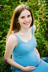 Fototapeta premium Pregnant happy girl sits and hold hands on stomach, sitting on background green grass in field in the outdoor. Close up. upper half. Looking at camera.