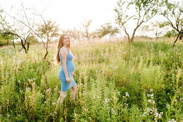 Pregnant happy girl go, with round stomach, go on grass in the outdoor in the garden background with trees. Close up. full length. go sideways. Looking at camera