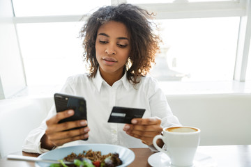 Portrait of young woman using cellphone and credit card in cafe