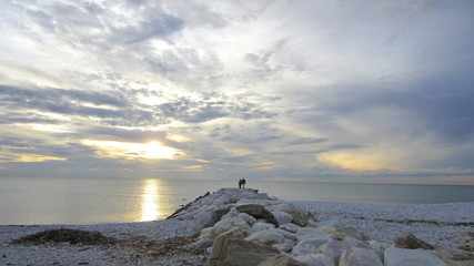 The couple watches the sunset over the sea, standing on a rocky waterfront. Cloudy pastel sky and romantic atmosphere