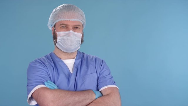 Dolly-in portrait shot of middle-aged male doctor wearing scrubs, gloves, bouffant cap and procedure mask standing isolated on blue background and posing for camera
