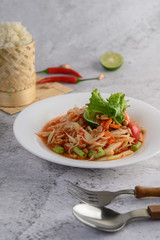 Thai papaya salad in a white plate with Sticky rice, chili, Spoon, and fork.