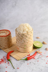Thai sticky rice in a woven bamboo basket on a wooden panel with chilies, lime, and garlic.