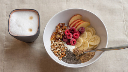 Healthy breakfast bowl with chia seeds, nuts, dates, raspberries, bananas, apples and yogurt with spoon in it. There is a cup of cappuccino near the bowl. The bowl is standing on linen napkin
