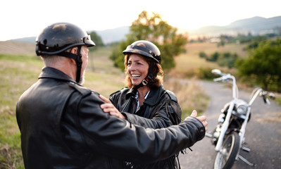 Cheerful senior couple travellers with motorbike in countryside, talking.