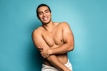 Fototapeta na wymiar Beauty portrait of half naked handsome smiling young man with beautiful torso dressed in towel touching his biceps looking at camera isolated over blue background